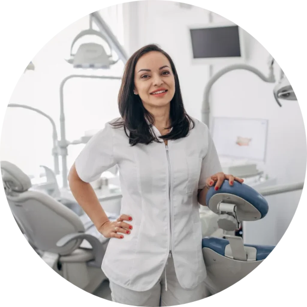 Una mujer con uniforme dental blanco junto a un sill&oacute;n dental, sonriendo, con herramientas NSK visibles. | La muela